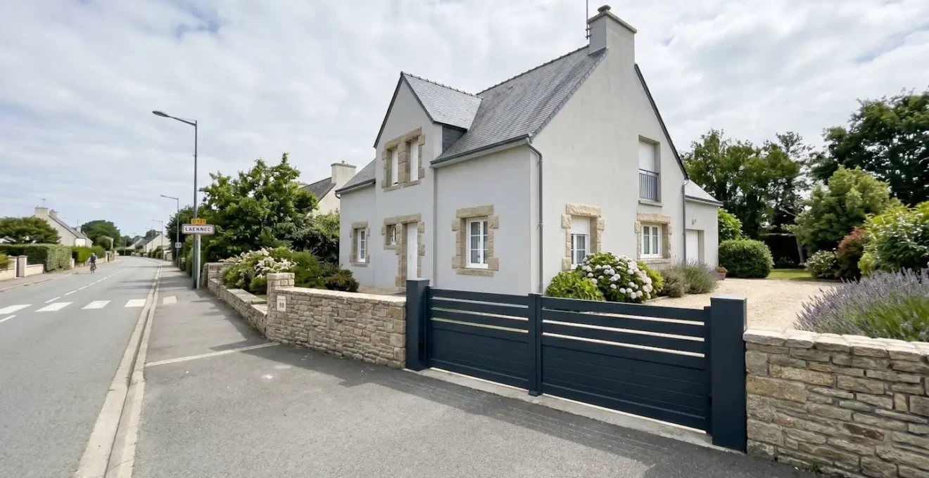 Un portail coulissant en aluminium anthracite fermé devant une maison moderne à façade en enduit gris clair avec des encadrements de fenêtres en pierre, sous un ciel lumineux de Bretagne